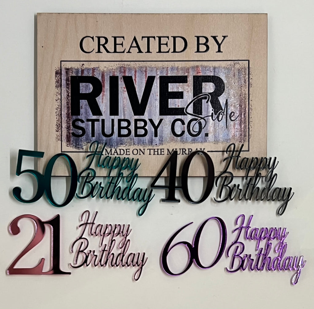 Mirror acrylic Happy Birthday milestone number charms including 21, 40, 50 and 60 in assorted mirror colours arranged below a Riverside Stubby Co wooden sign