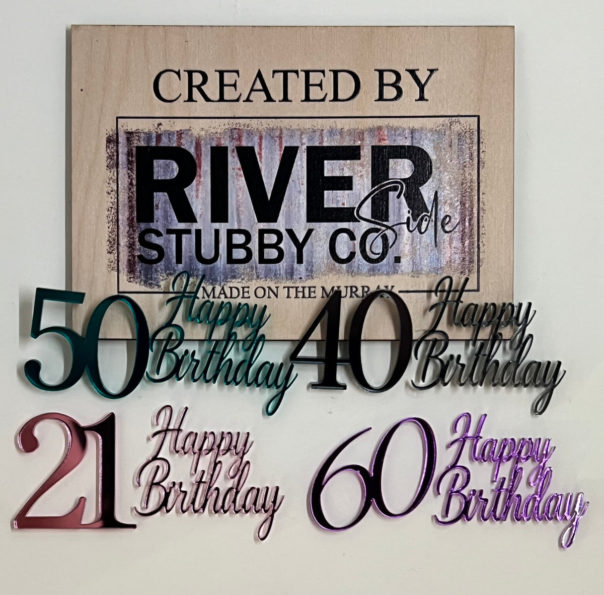 Mirror acrylic Happy Birthday milestone number charms including 21, 40, 50 and 60 in assorted mirror colours arranged below a Riverside Stubby Co wooden sign